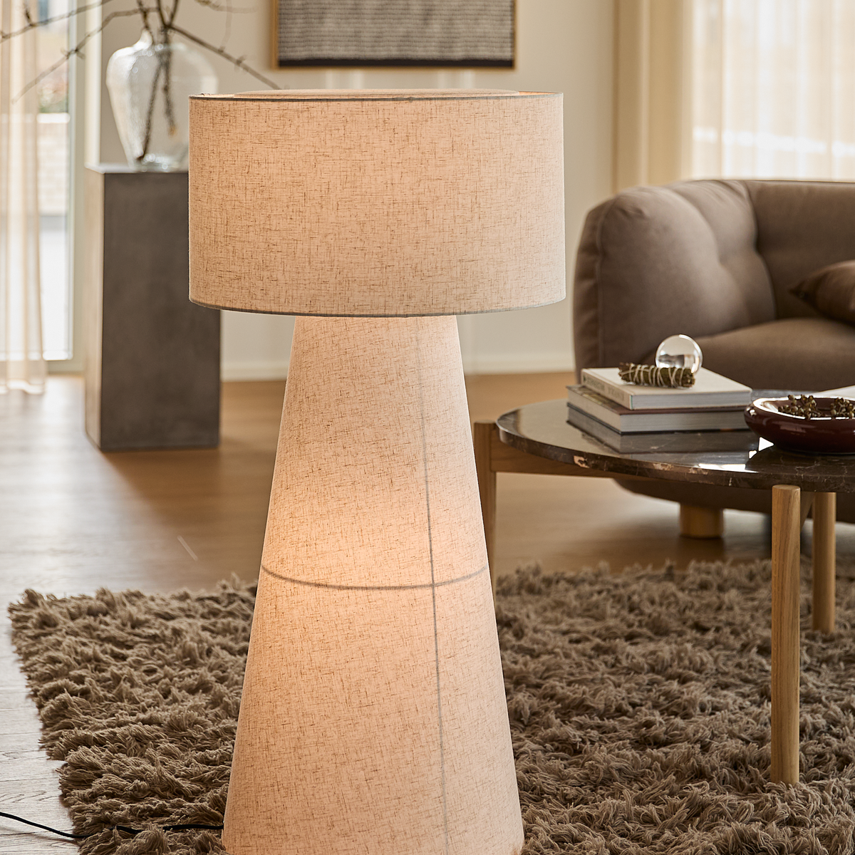 Stor gulvlampe i beige stof i en stue med blødt ryatæppe, sofabord med bøger og en sofa.