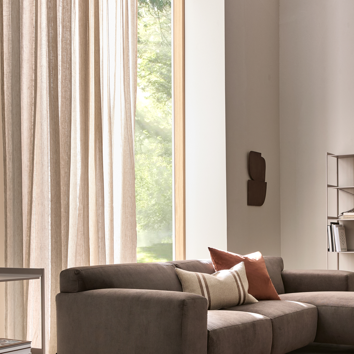 Lyst stue med grå modulsofa, lyse, lette gardiner ved stort vindue og puder i terrakotta og beige.