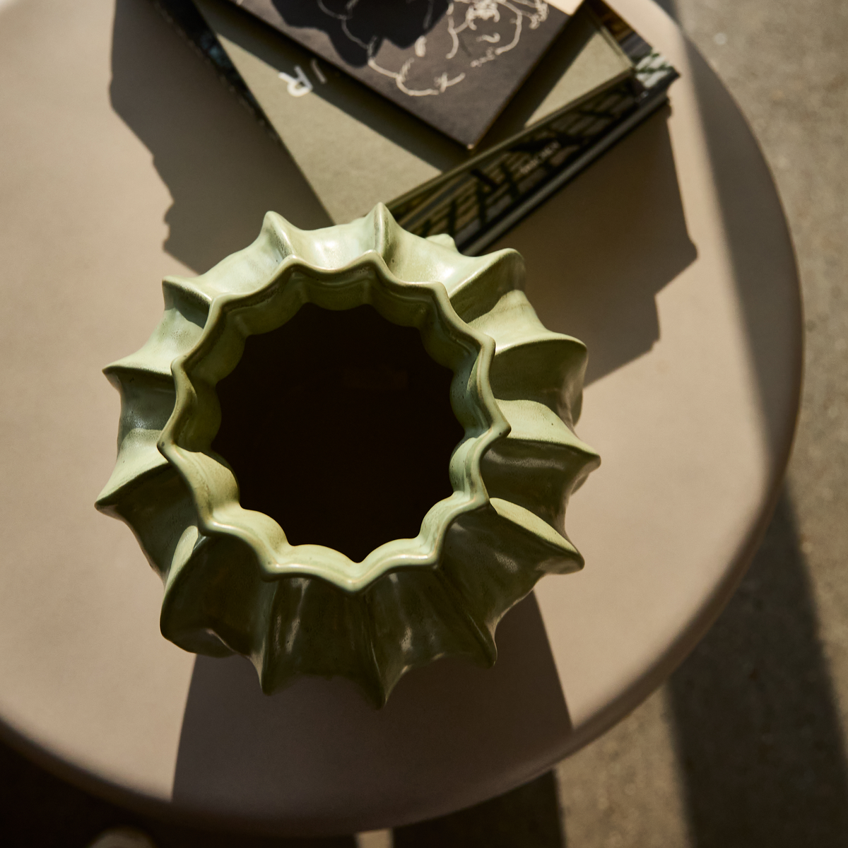 Grøn skulpturel vase med bølget kant på rund bordplade i sollys, med stablede bøger ved siden af.