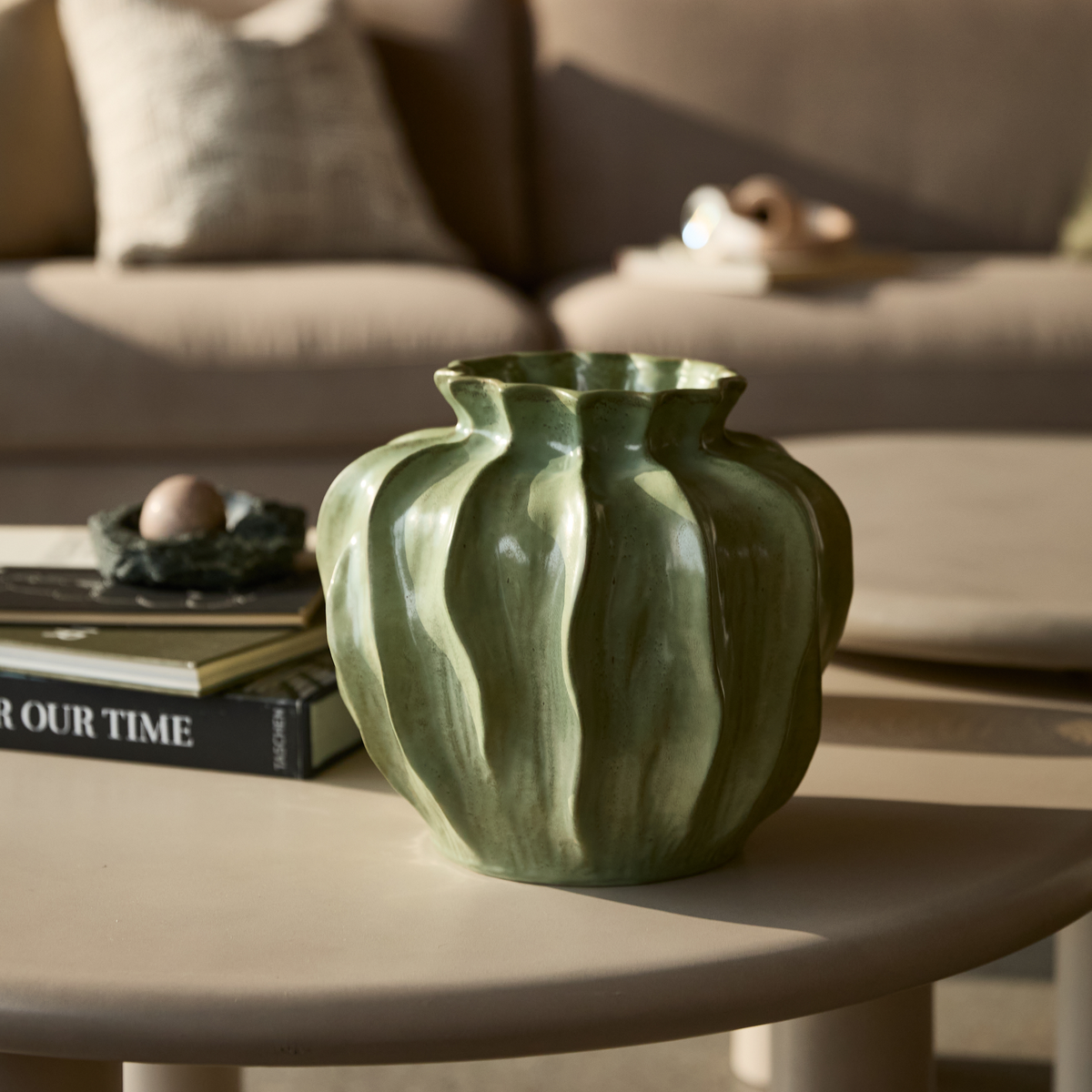 Grøn bølget keramisk vase på beige sofabord ved siden af bøger, bl.a. “OUR TIME”, i en varm stue.
