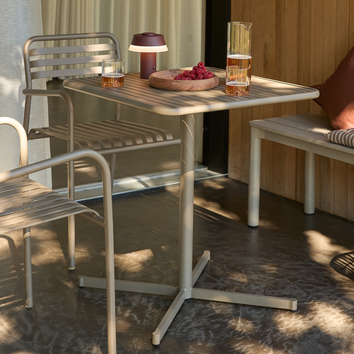 Beige havemøbler på en terrasse: et metalbord med to stole, bænk med pude, en lille bærbar bordlampe, glas og karaffel med drik samt en trætallerken med hindbær.