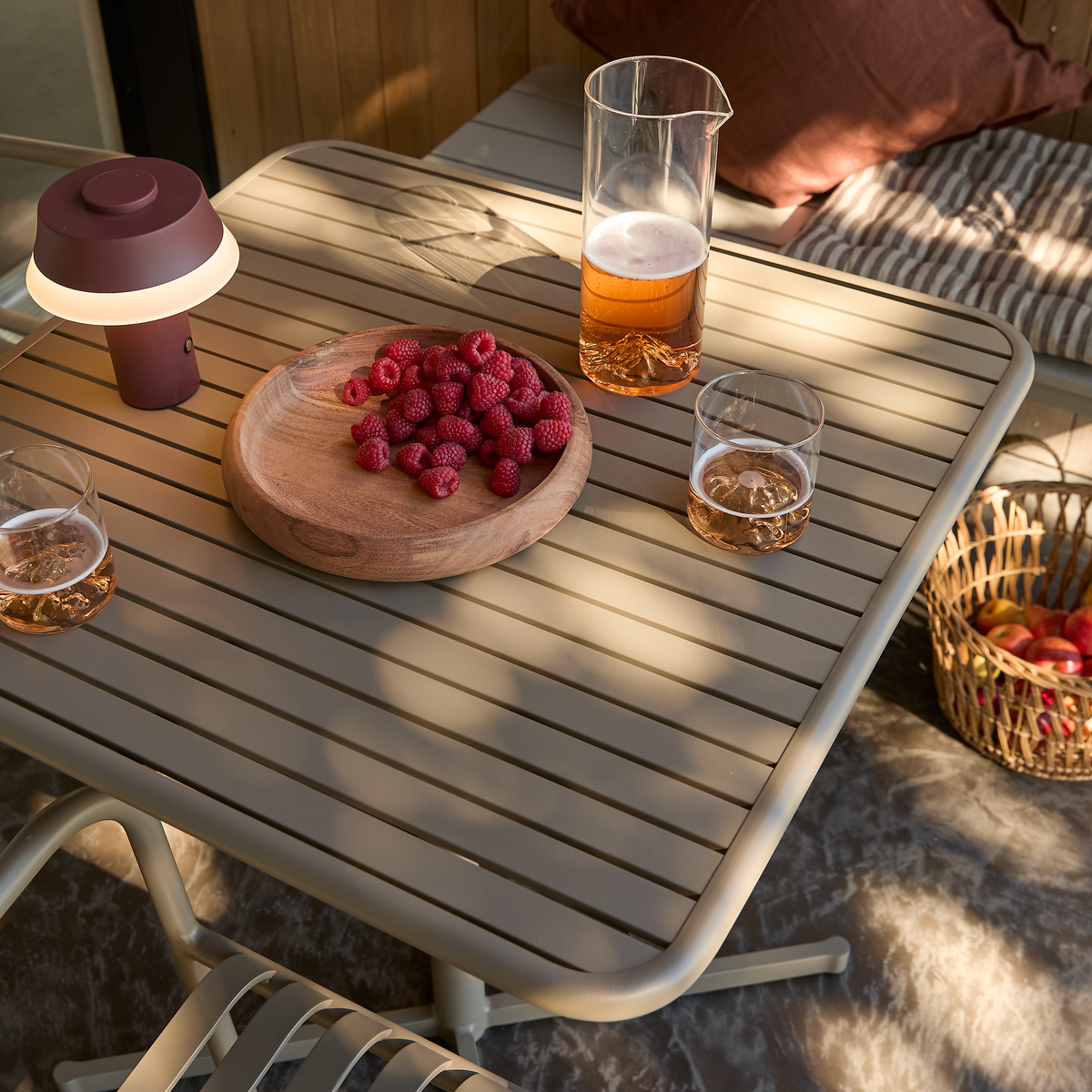 Terrassebord med træfad med hindbær, kande med roséfarvet drik, to glas, bærbar bordlampe og kurv med æbler i varmt sollys.