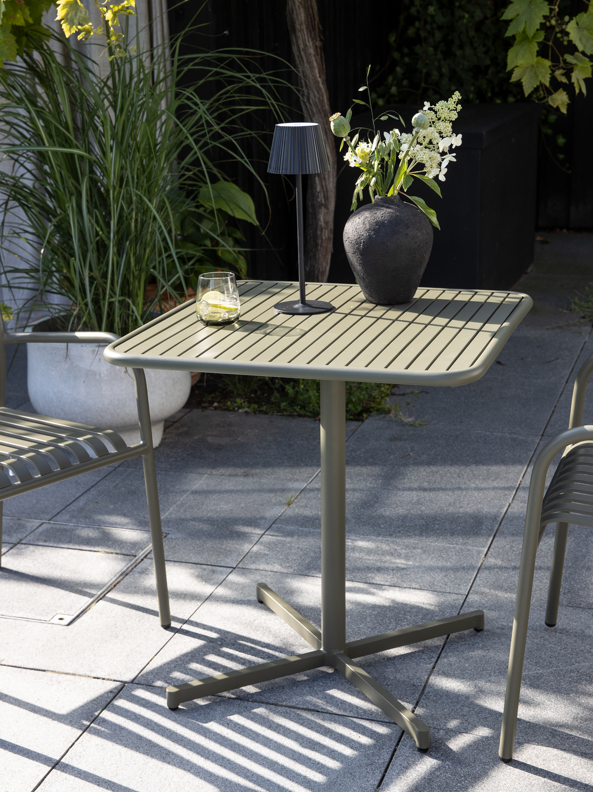 Olivengrønt udendørsbord med rillet bordplade, lille sort bordlampe, vase med hvide blomster og et glas med citron på en grå terrasse med matchende stole og planter.