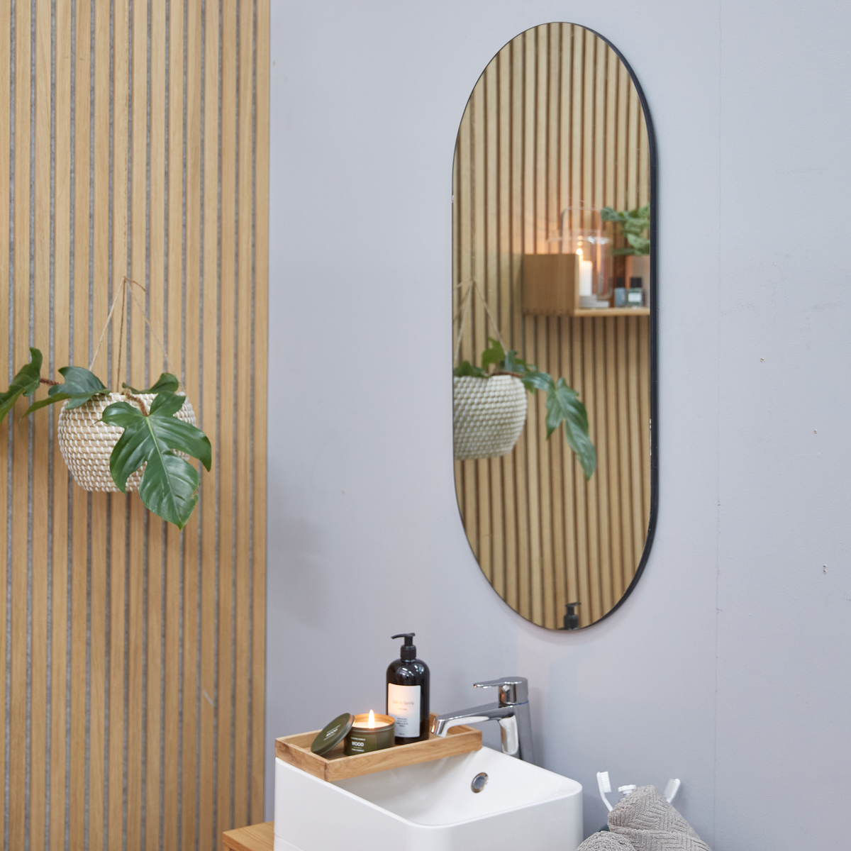 Minimalistisk badeværelsesscene med oval spejl på grå væg ved siden af trælameller, lille vask med træbakke med tændt duftlys og sæbedispenser samt en hængende plante med grønne blade.