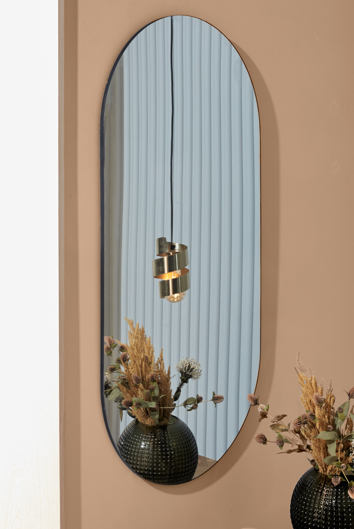 Oval vægspejl på beige væg, der spejler en plisseret lyseblå gardin, en messingpendel og en sort struktureret vase med tørrede blomster.