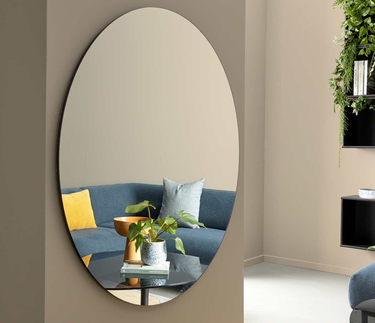 Oval vægspejl, der afspejler en blå sofa med gul pude, sofabord med plante og bøger samt beige vægge og hængende grønne planter.
