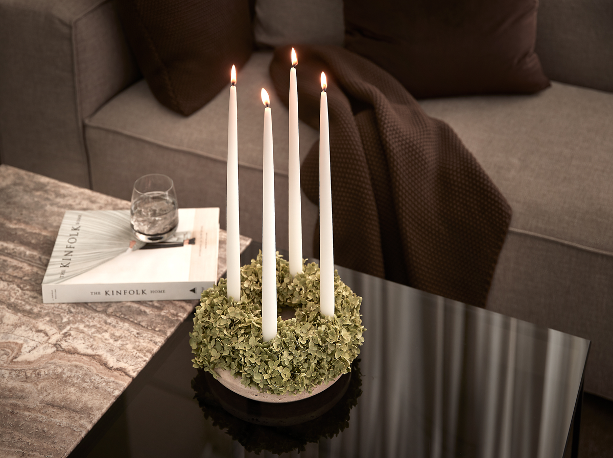Fire tændte hvide kronelys i en grøn krans som centerpiece på et sort glasbord i stuen; ved siden af ligger en bog med titlen “THE KINFOLK HOME” og et glas vand, med grå sofa og brun plaid i baggrunden.