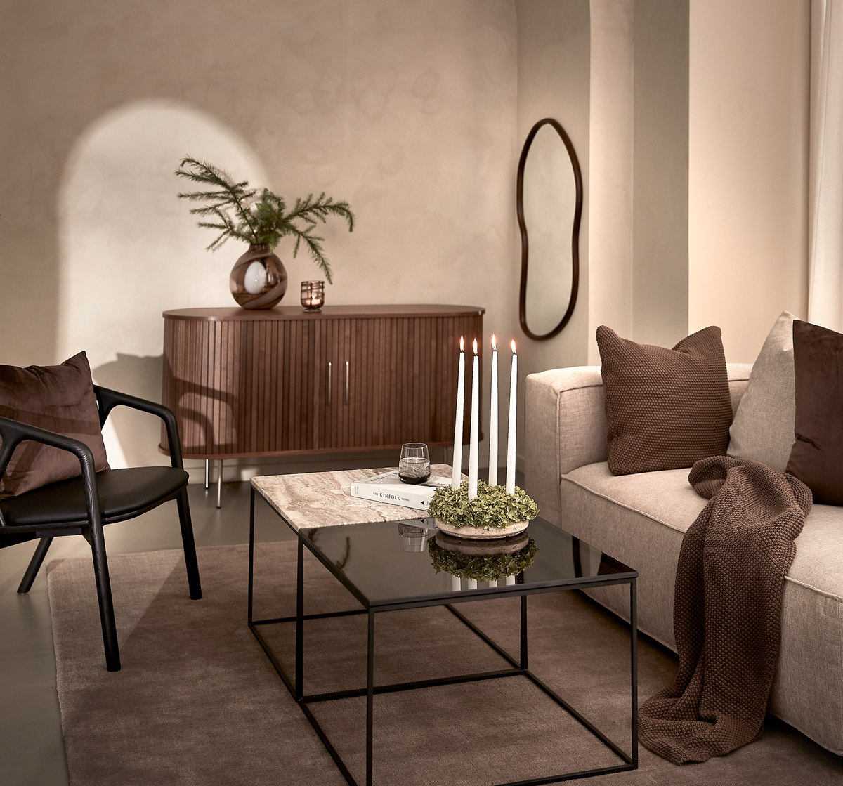 Varm, minimalistisk stue med beige sofa, brune puder og plaid, sort stol og glas-/sten-sofabord med fire tændte lys og bøger; træskænk med vase med grønne grene og organisk spejl på væggen.