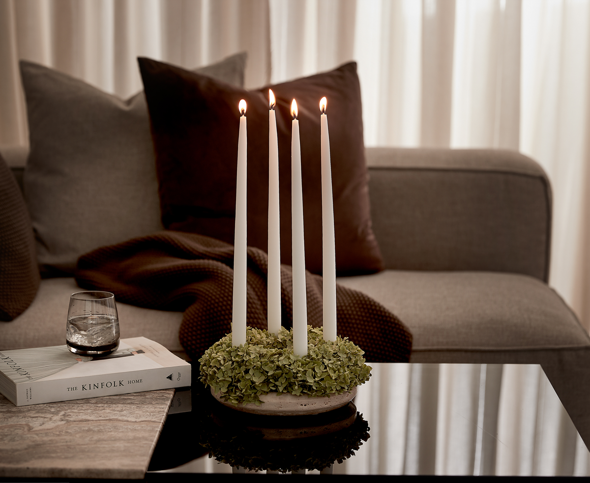 Fire tændte hvide stagelys i en grøn krans på sofabordet, med et glas og bogen ”THE KINFOLK HOME” foran en sofa med puder og brun plaid.