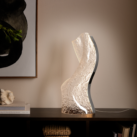 Skulpturel, bølgeformet bordlampe i struktureret glas, der lyser varmt på en mørk hylde, ved siden af en stak bøger (bl.a. VOL VII), en organisk sten og et abstrakt vægbillede.