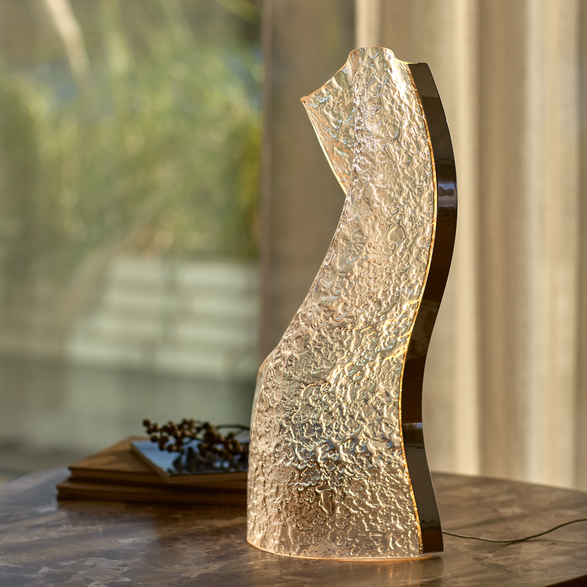 Skulpturel bordlampe i tekstureret glas med varm glød på et marmorbord, sløret baggrund og et par bøger.