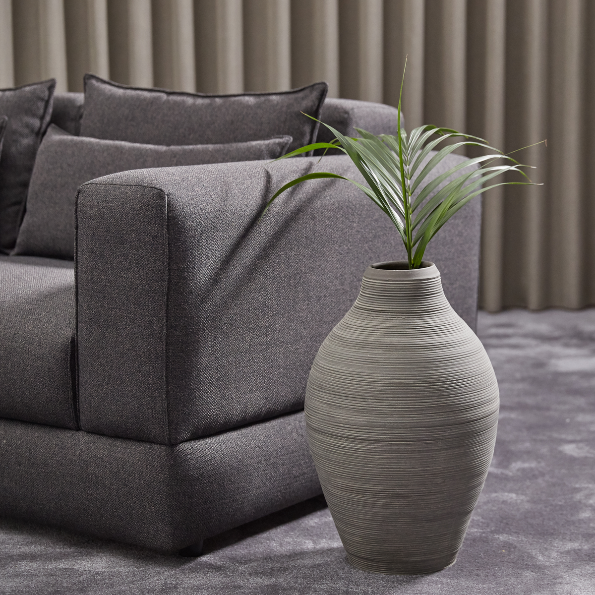 Grå sofa med puder og en stor riflet grå gulvvase med grøn plante på gråt tæppe foran beige gardiner.