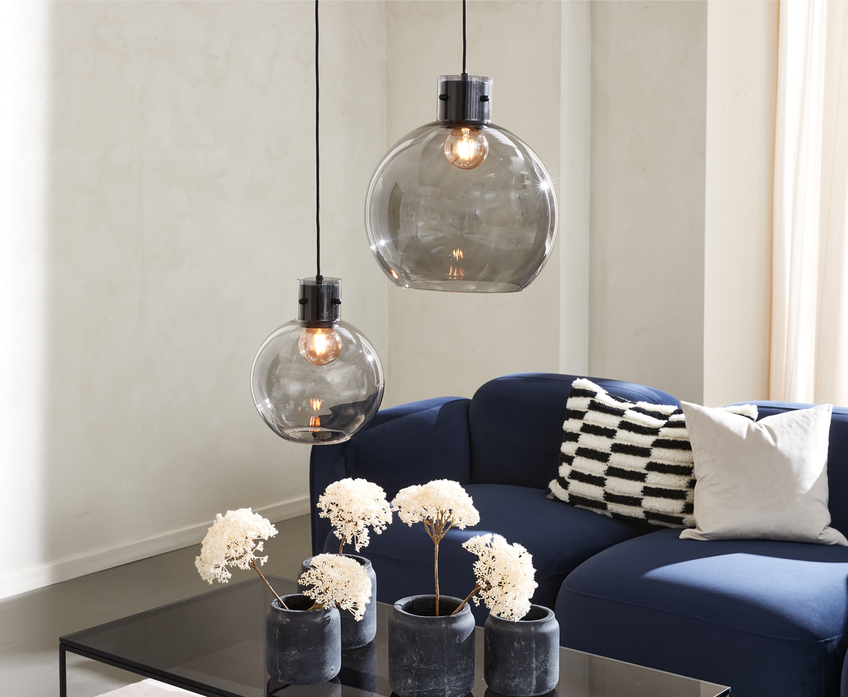 To røgfarvede glaspendler hænger over en blå sofa med sort-hvide puder og små sorte vaser med hvide tørrede blomster på et glas sofabord i en lys stue.