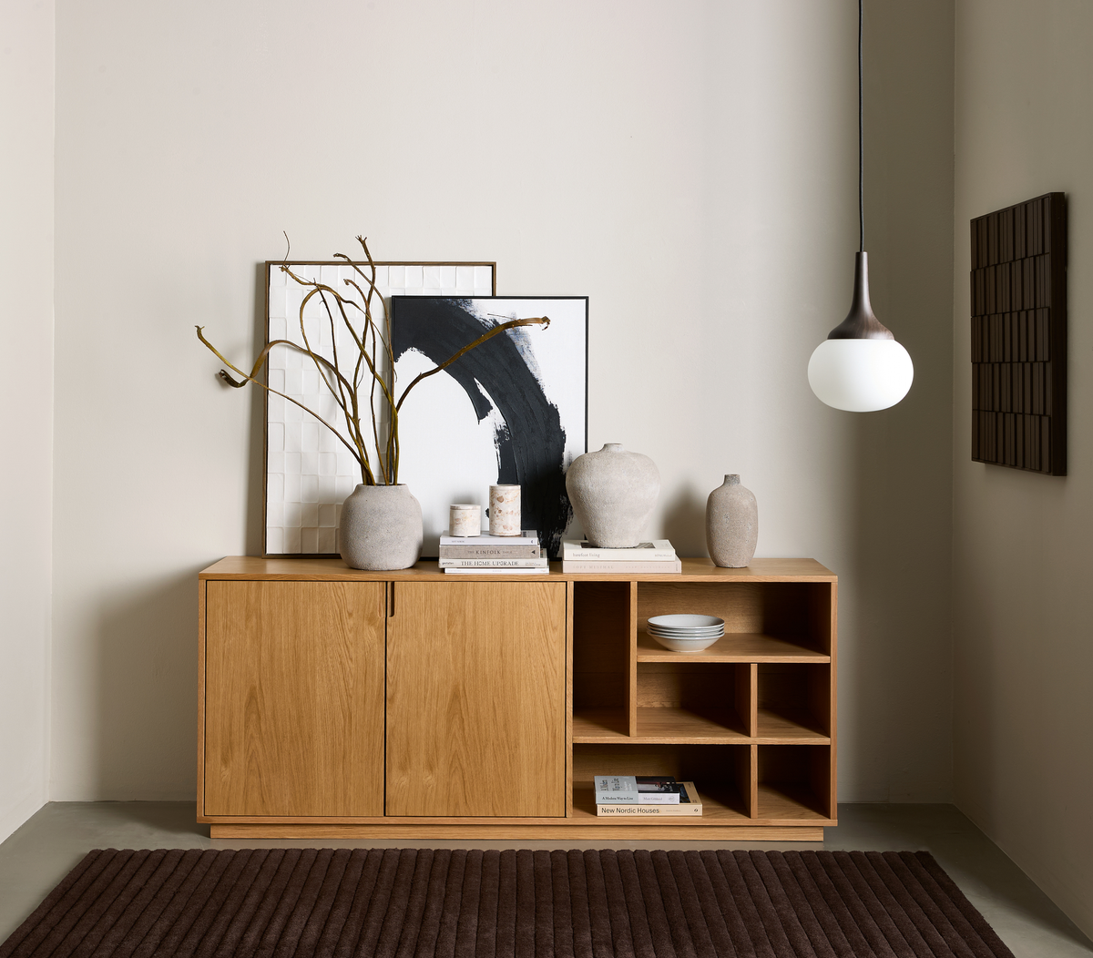 Minimalistisk krog med et skænk i egetræ, neutrale vaser, to indrammede kunstværker, en hængende kuglependel og bøger – en bog har titlen “New Nordic Houses”.