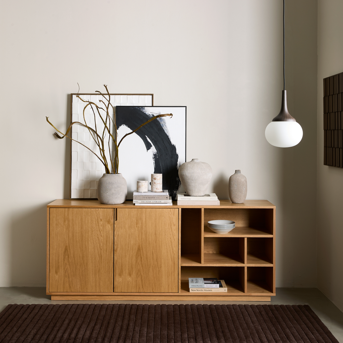 Minimalistisk scene med egetræsskænk, keramiske vaser, indrammet kunst, bøger inklusive “New Nordic Houses”, skåle på åbne hylder, hængende pendellampe og brun tæppe.