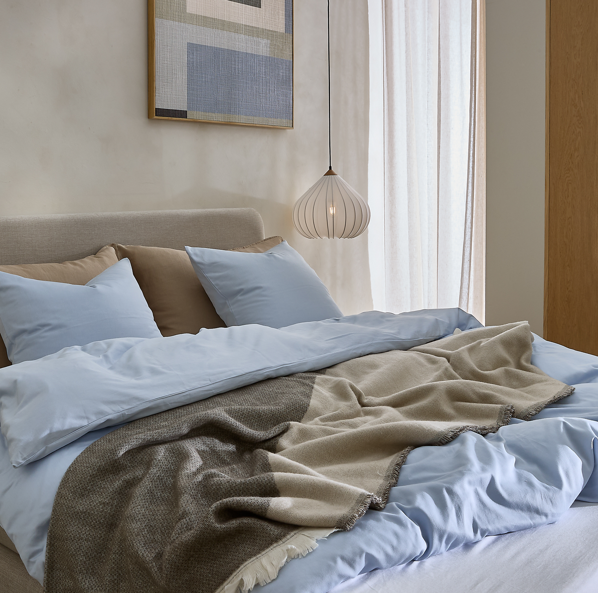 Lyst soveværelse med redt seng i lyseblåt sengesæt, beige puder og en beige‑grå plaid, hængende hvid pendellampe, gardiner og abstrakt vægkunst i neutrale toner.