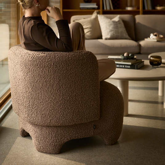 Brun bouclé-lænestol med person i profil i en varm stue; sofabord med bøger inklusive titlen ”Our Time” og beige sofa med puder i baggrunden.