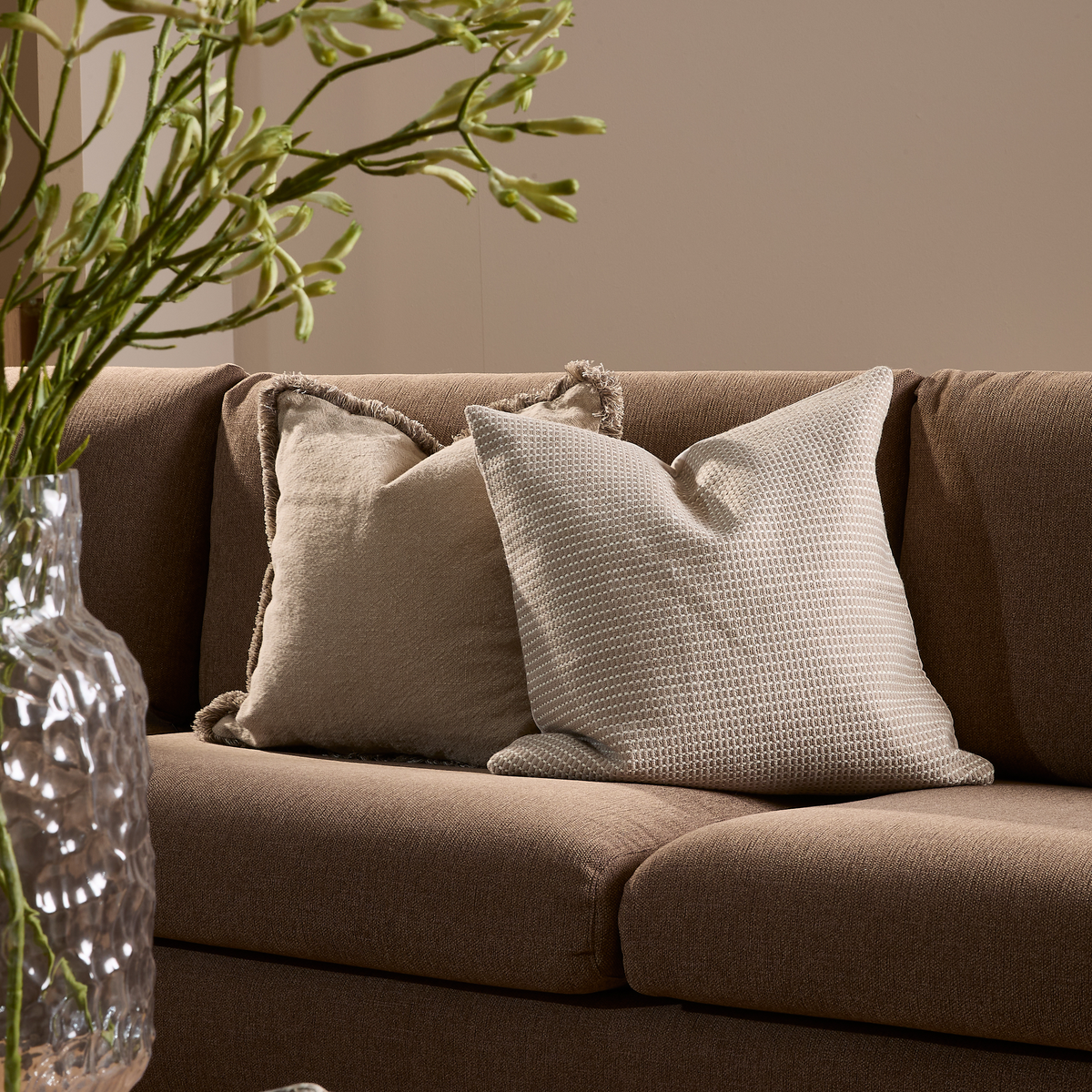 To beige puder på en brun sofa med en glasvase med grønne grene i forgrunden i en rolig stue.