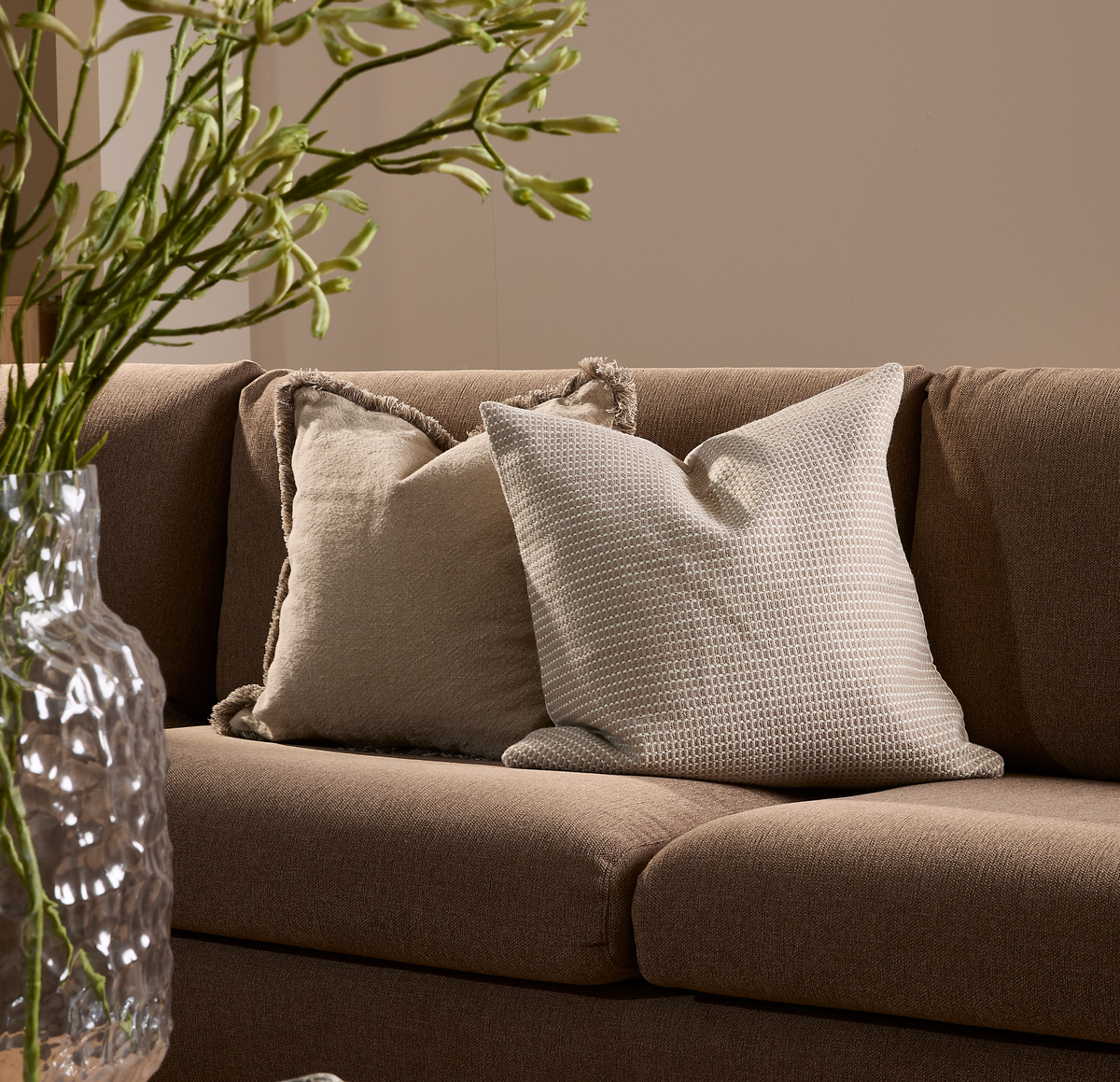Brun sofa med to beige pyntepuder og en glasvase med grønne grene i forgrunden.