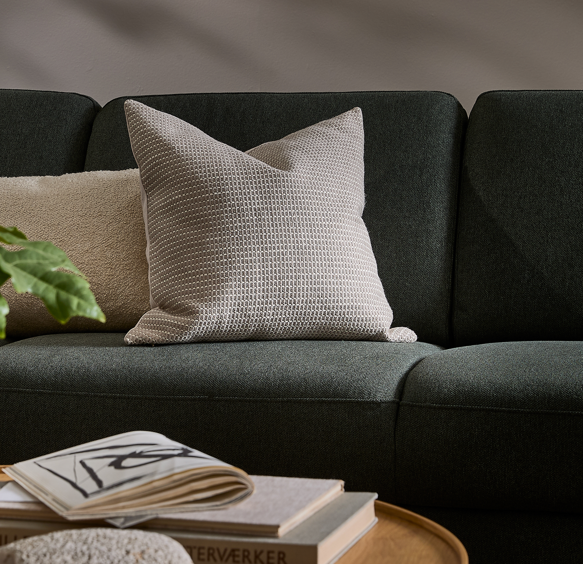 Mørkegrøn sofa med beige, tekstureret pyntepude; bøger ligger på et rundt sofabord i blødt dagslys for en rolig, skandinavisk stemning.