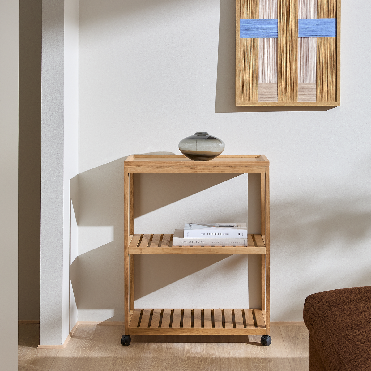 Minimalistisk reol i lyst egetræ på små hjul med lamelhylder; lille keramikvase ovenpå, vægbillede med blå detaljer og bøgerne Soft Minimal og The Kinfolk Home på midterhylden i en lys stue med sollys.