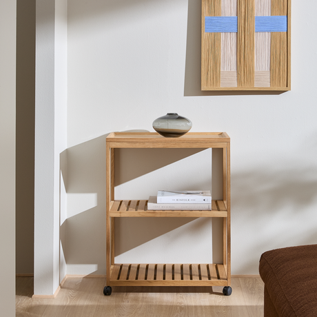 Minimalistisk reol i lyst egetræ på små hjul med lamelhylder; lille keramikvase ovenpå, vægbillede med blå detaljer og bøgerne Soft Minimal og The Kinfolk Home på midterhylden i en lys stue med sollys.