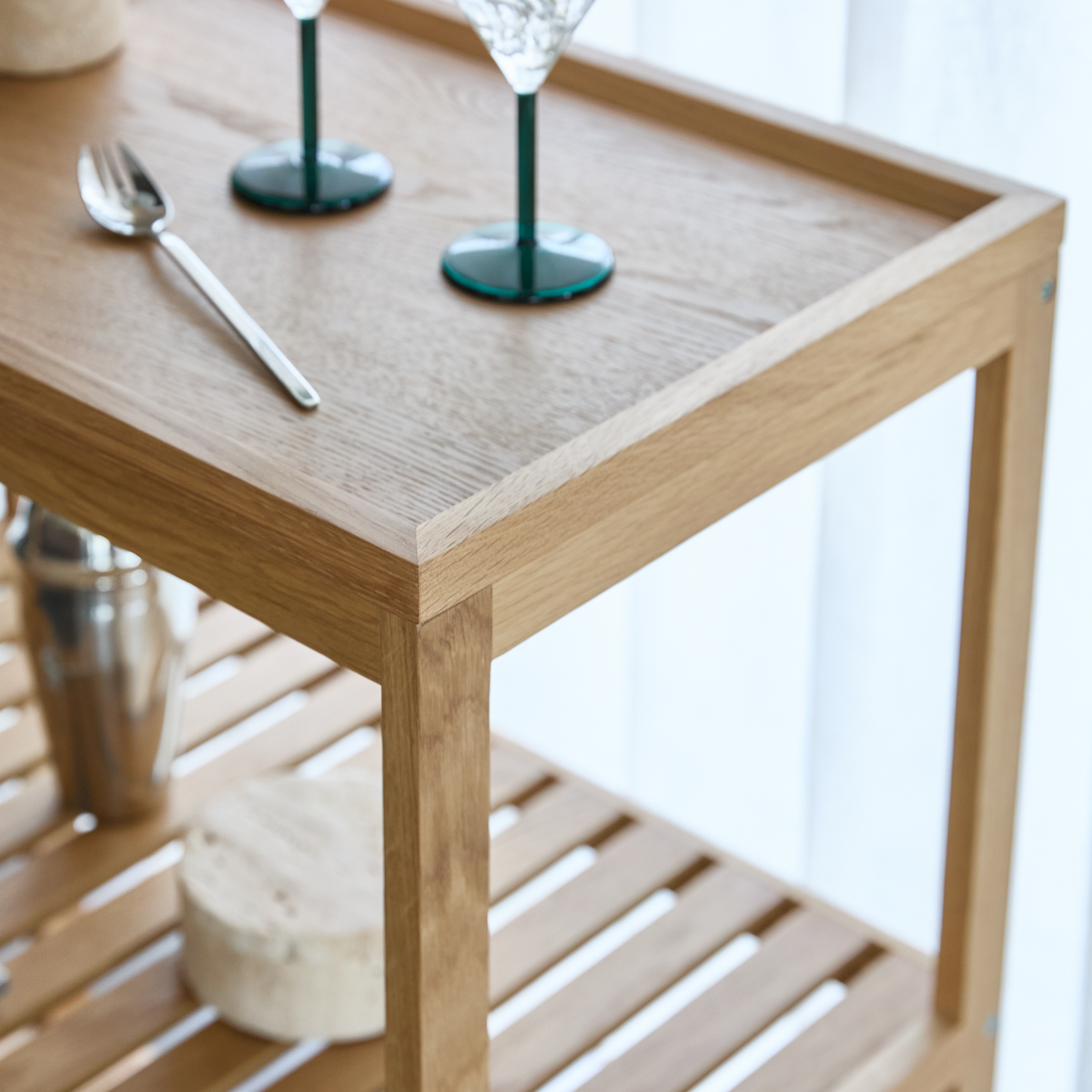 Nærbillede af et lyst bakke­bord/serveringsvogn i træ med lamelhylde, to glas med grøn fod og en ske i blødt dagslys ved en hvid gardin.