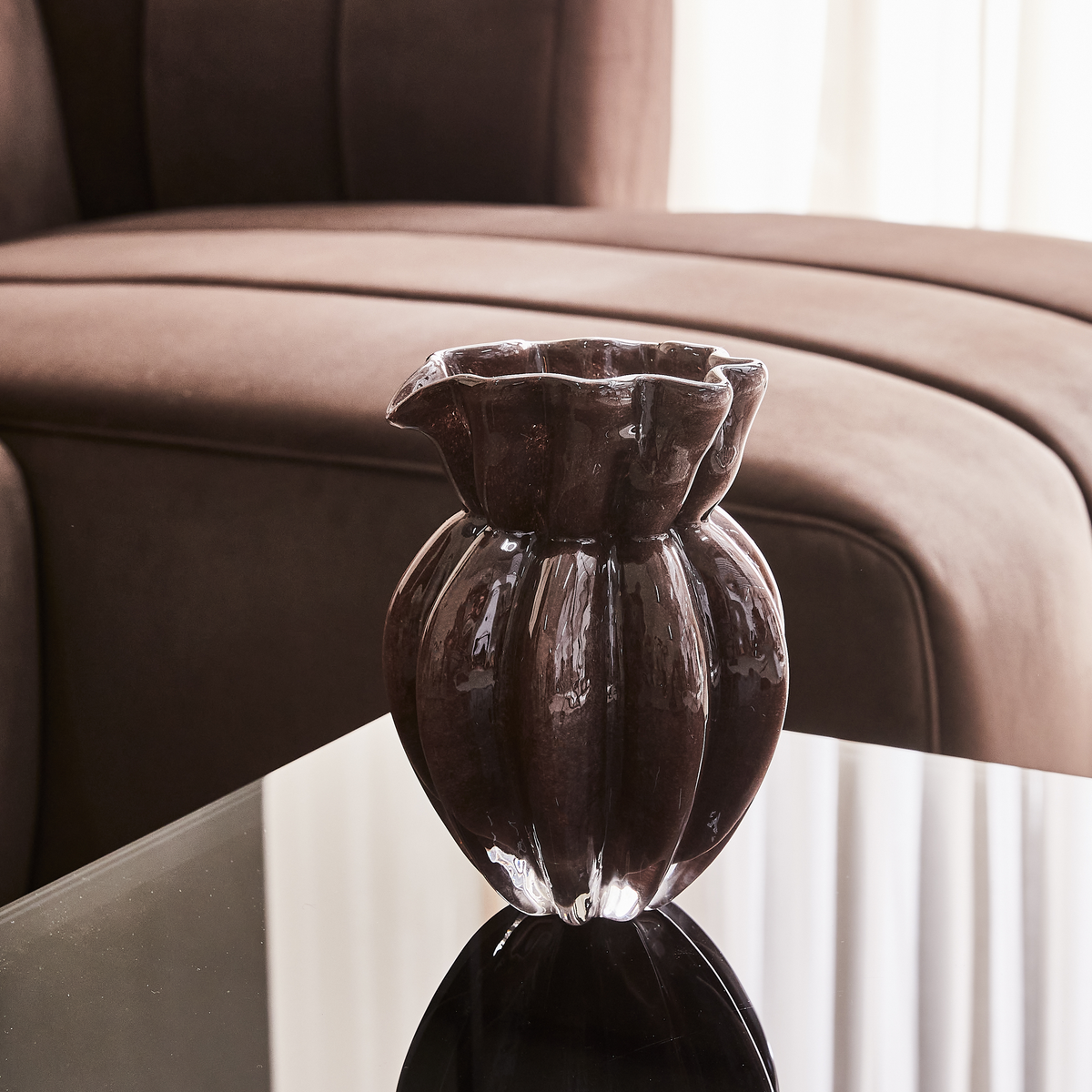 Mørkebrun glaseret vase med bølget kant på sort glasbord foran brun sofa i dagslys.