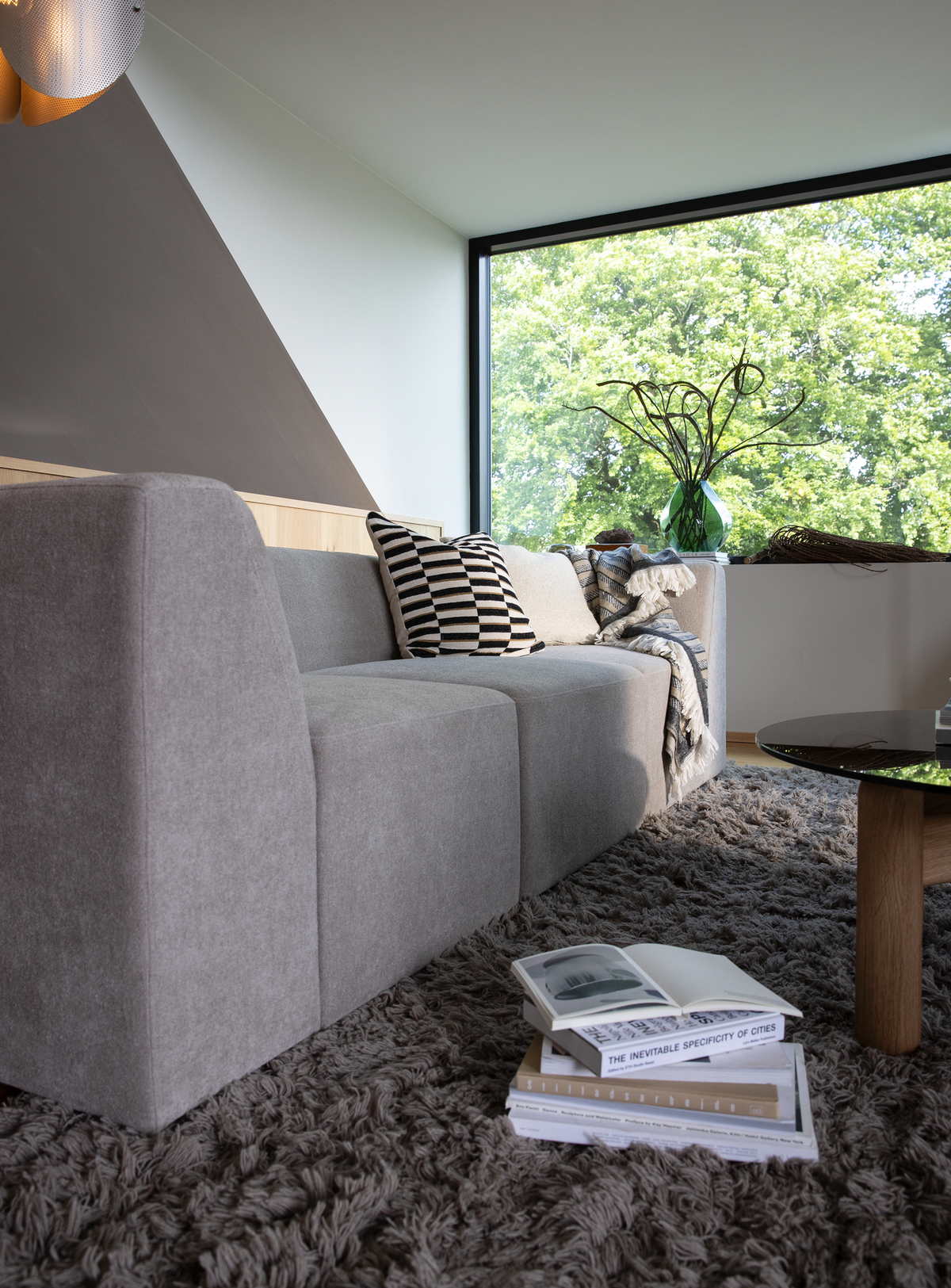 Moderne stue med grå modulsofa, stribet pude og plaid på en langhåret tæppe; en stak bøger på gulvet med “The Inevitable Specificity of Cities”; rundt glasbord og stort vindue med grøn vase og grene mod grønne træer.