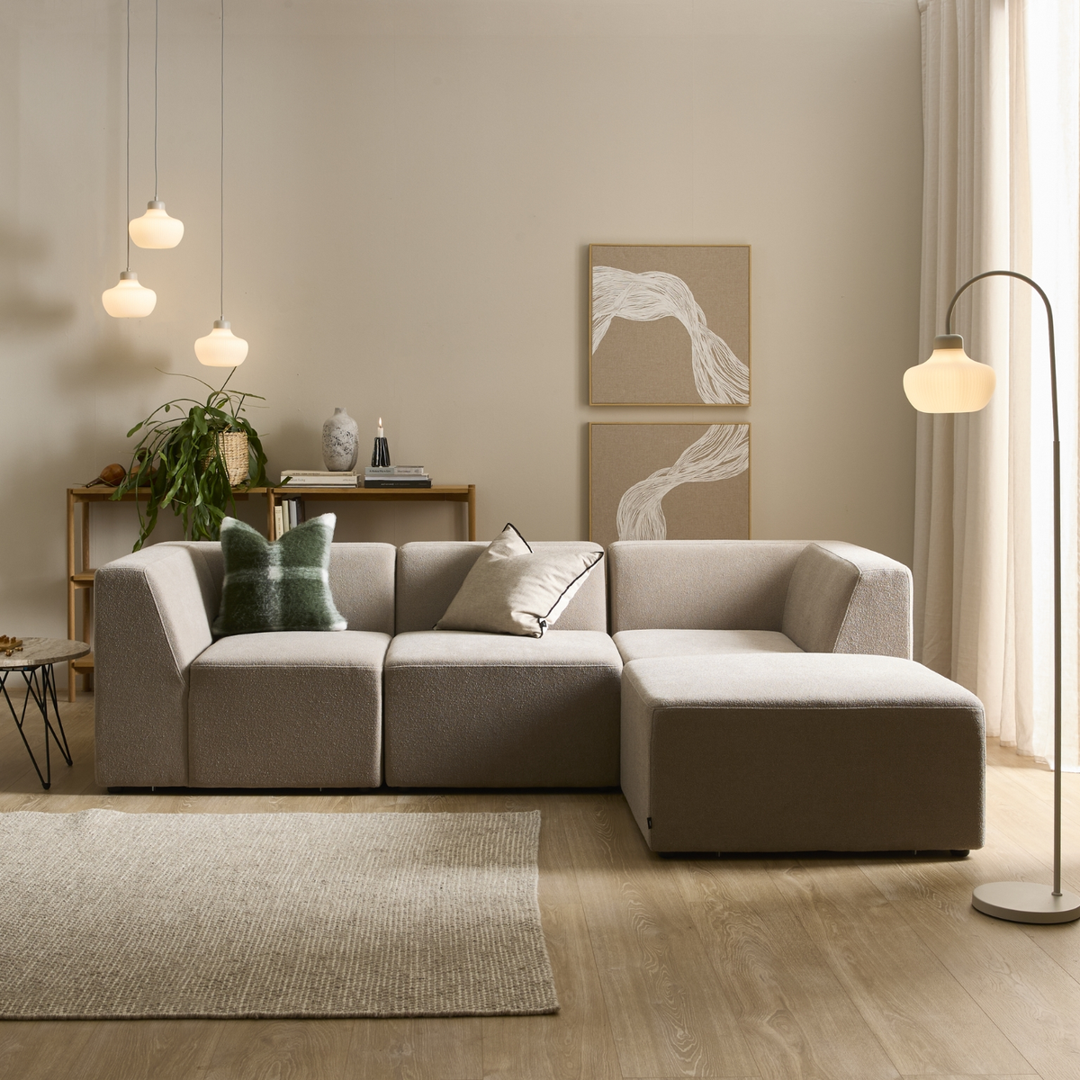 Beige modulsofa i lys skandinavisk stue med gulvlampe, tre pendler, vægkunst, tæppe og grøn plante.