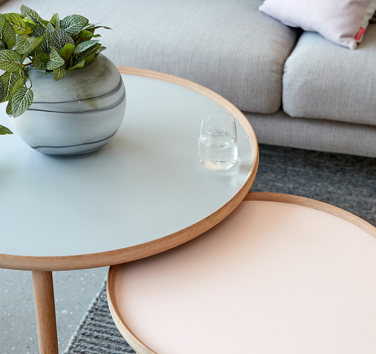 Nærbillede af to runde sofaborde i sæt med lyseblå og pudderrosa plader og trækanter, med et lille glas vand og en rund vase med grøn plante ved siden af en lysegrå sofa.