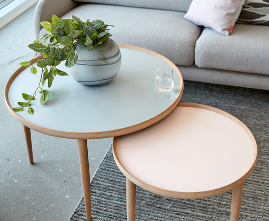 To runde sideborde i lyse pasteller med kant i træ; en vase med grønne blade og et glas vand på det større bord, grå sofa og tæppe i baggrunden.