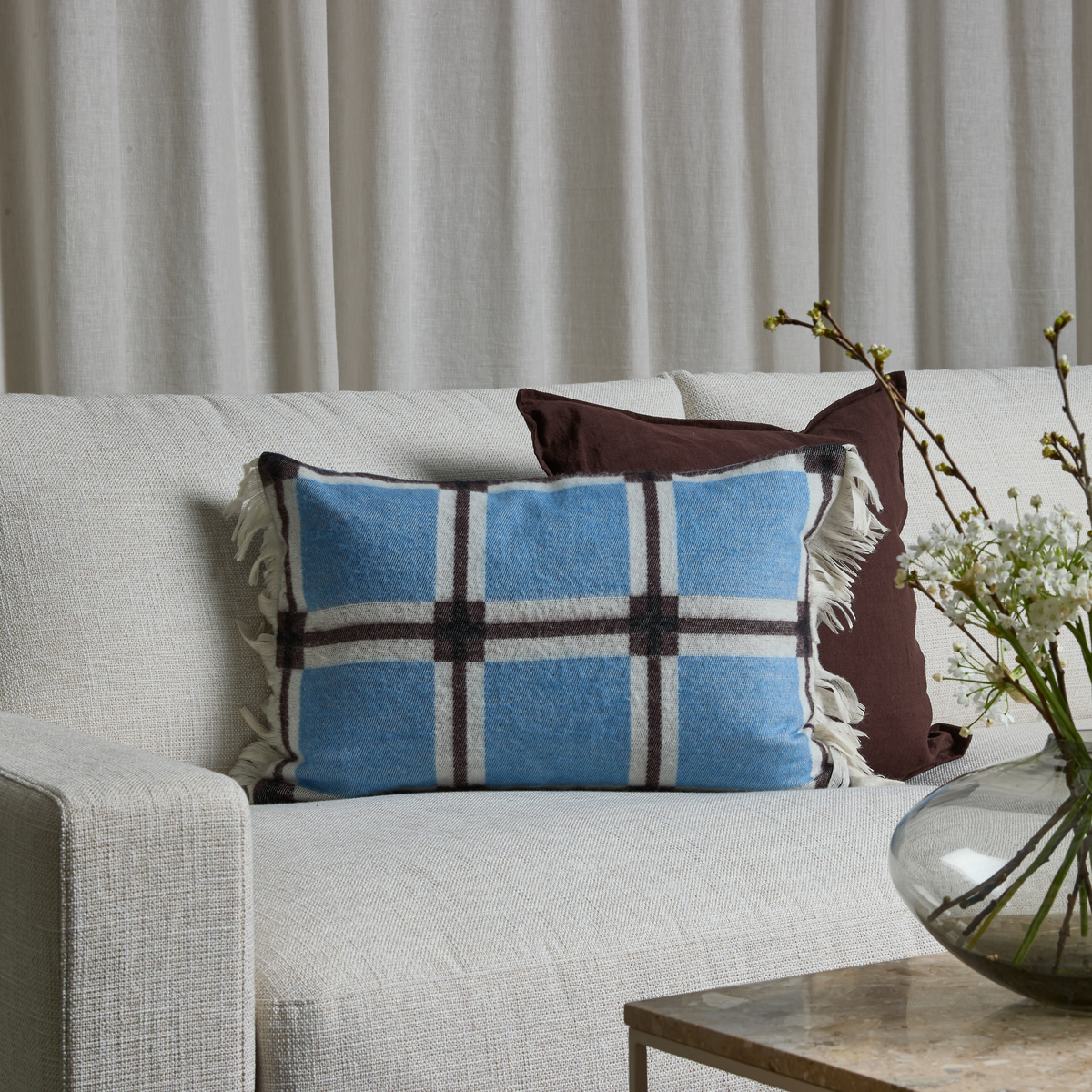 Hvid sofa med blå ternet pude med frynser, brun pude bagved og glasvase med hvide blomstergrene på bordet foran, mod lyse gardiner.