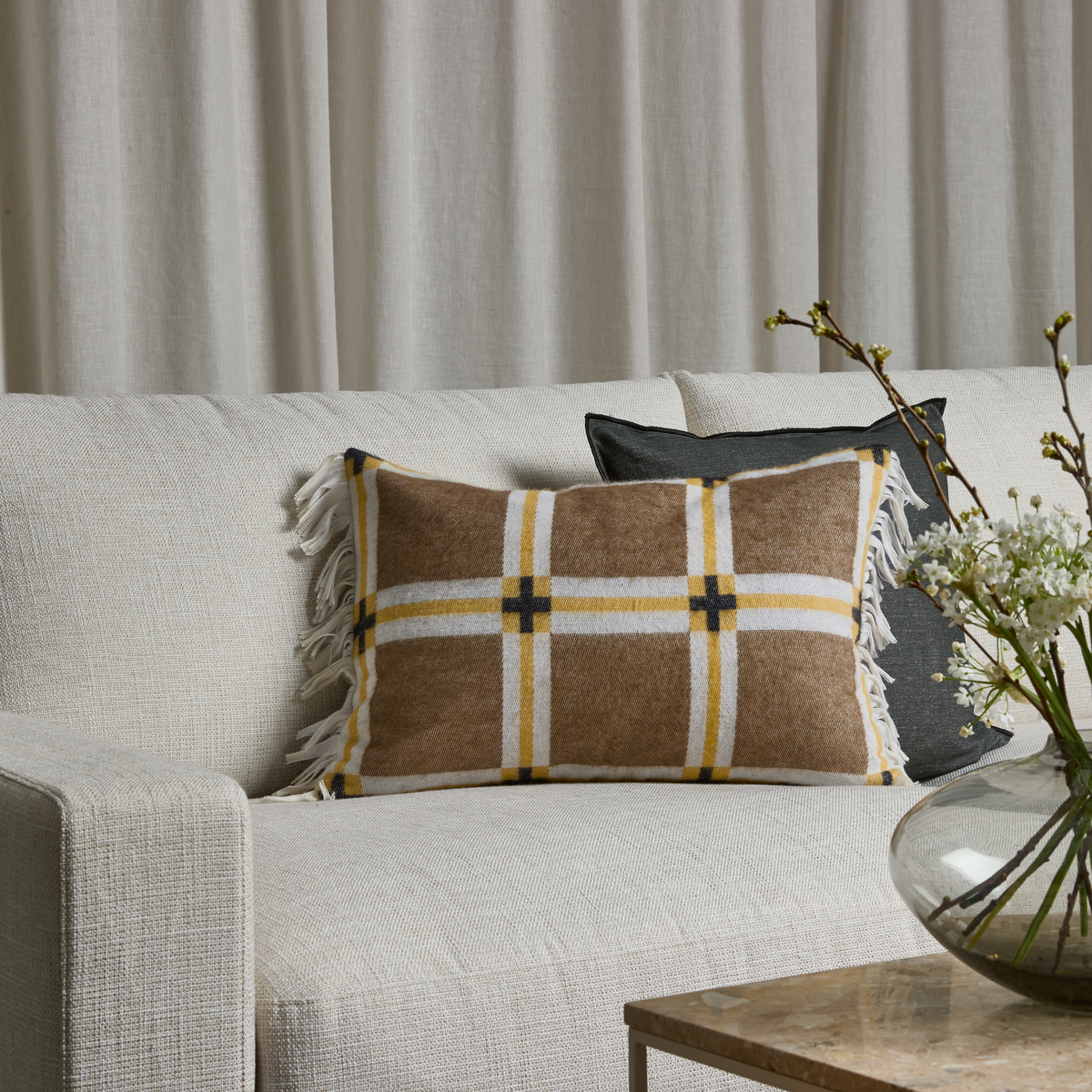 Beige sofa med rektangulær ternet pude i brun, hvid og gul med frynser, mørkegrå pude bagved og glasvase med hvide blomster på bordet foran lyse gardiner.