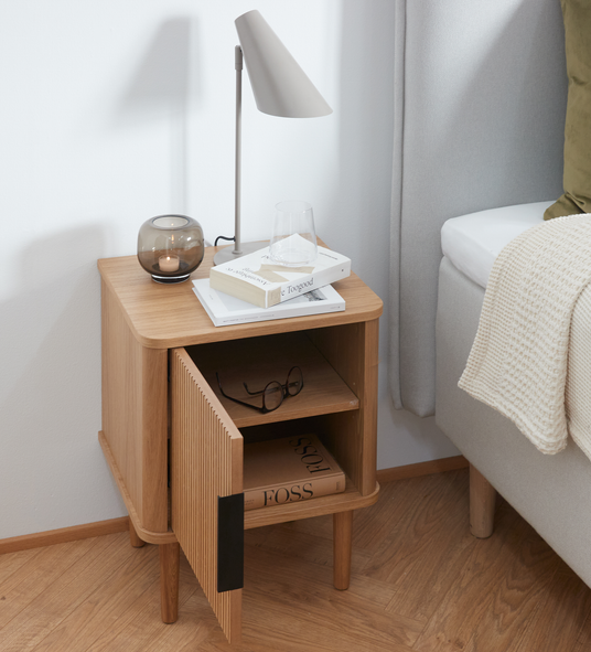 Lyst egetræsnatbord med rillet låge på klem, bordlampe, røgfarvet fyrfadsstage, drikkeglas og stablede bøger med titlerne ”Toogood” og ”FOSS” i et neutralt soveværelse med strikket plaid på sengen.