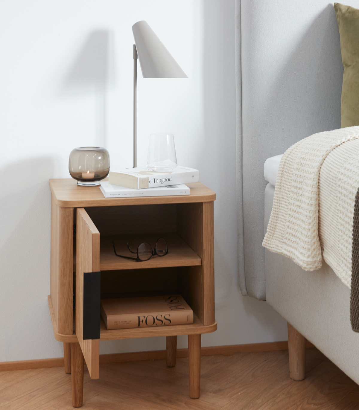 Minimalistisk soveværelsesscene med natbord i eg, bordlampe, glas og bøger Faye Toogood og FOSS; lågen står på klem ved siden af seng med plaid og pude.