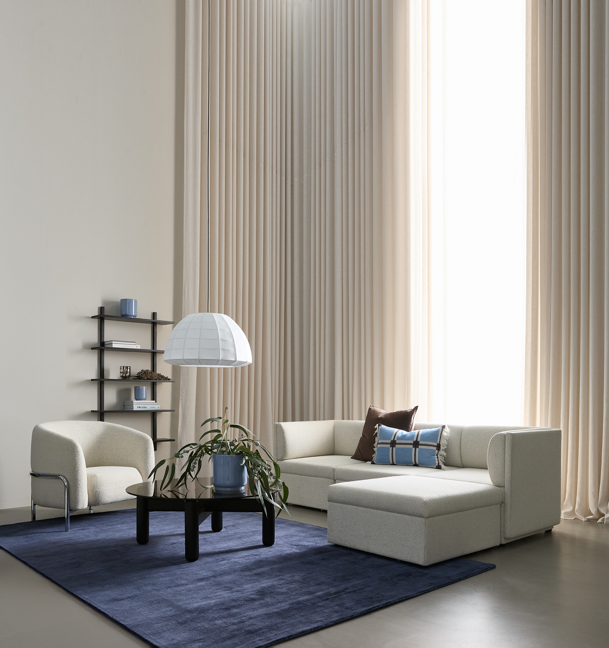 Lyst stue med beige modulsofa, blå tæppe, sort sofabord med grøn plante, pendel og høje beige gardiner.