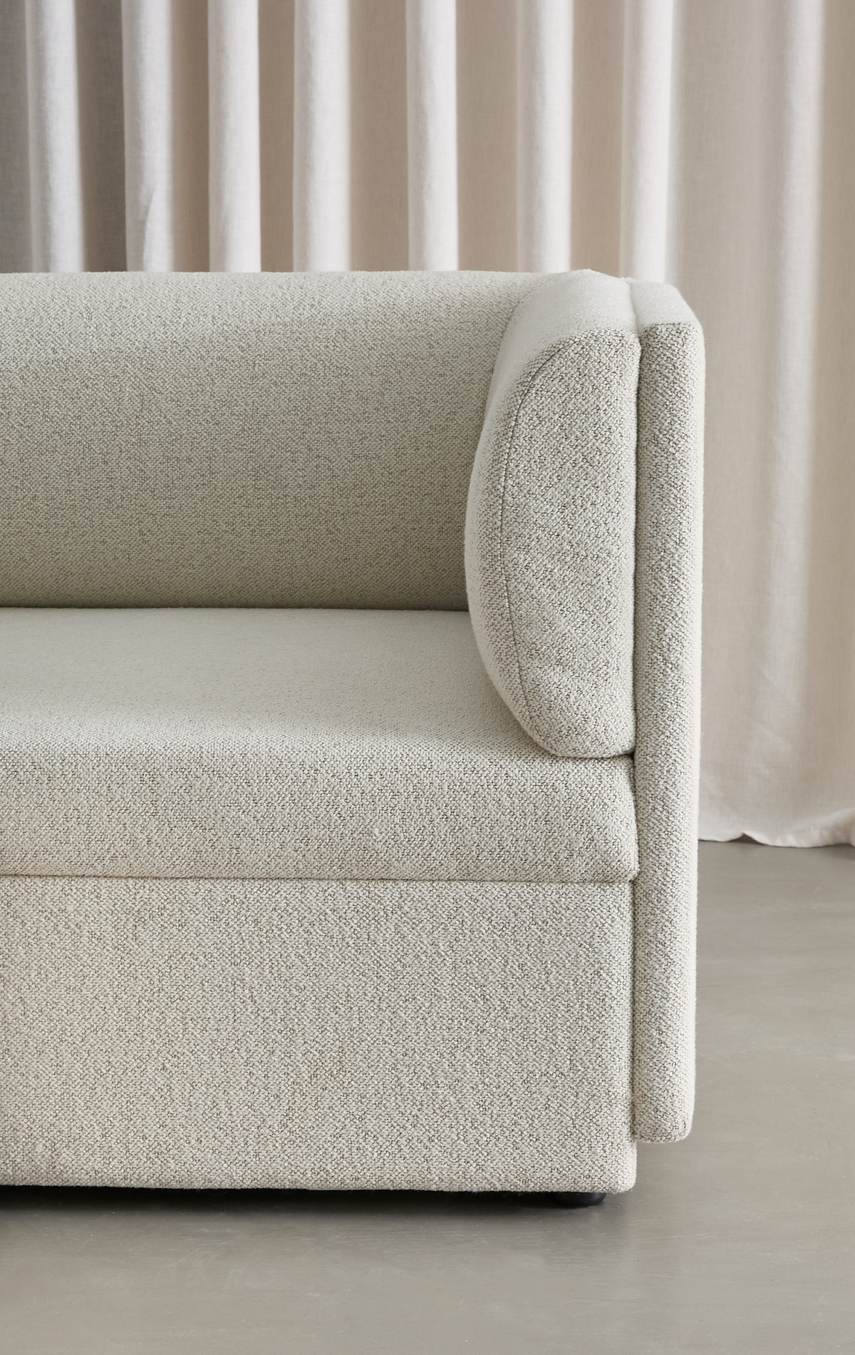 Nærbillede af en beige bouclésovesofa med afrundet armlænspude foran bløde beige gardiner og betongulv.