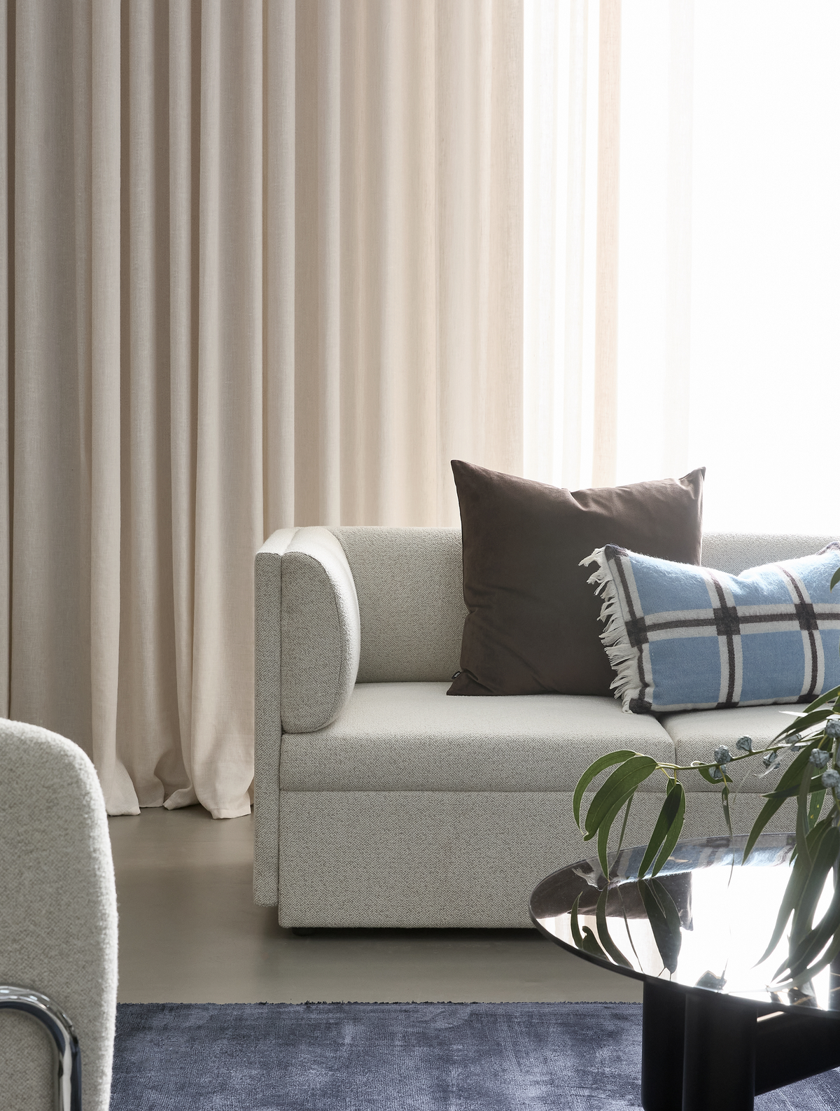 Lyst moderne stuehjørne med beige gardiner, lys bouclé-sofa med brun pude og blå ternet pude, glas sofabord med grønne kviste og blå tæppe.