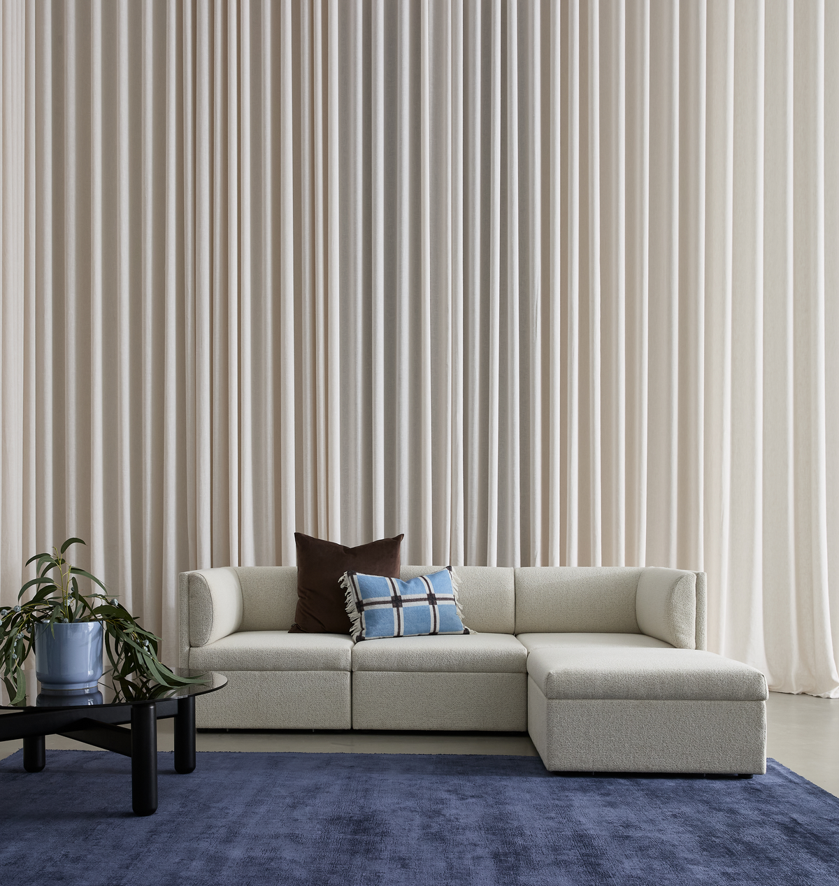 Lys beige modulsofa med chaiselong og to puder (brun og blå med tern) på et blåt tæppe, sort sofabord med grøn plante i blå potte foran gulvlange beige gardiner.