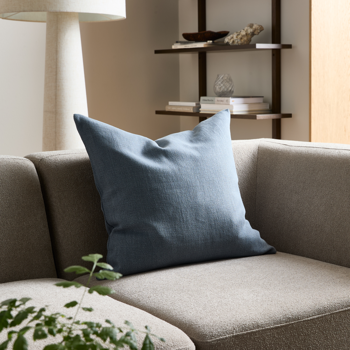 Beige sofa med blå pyntepude i stuen, gulvlampe og væghylder i baggrunden.