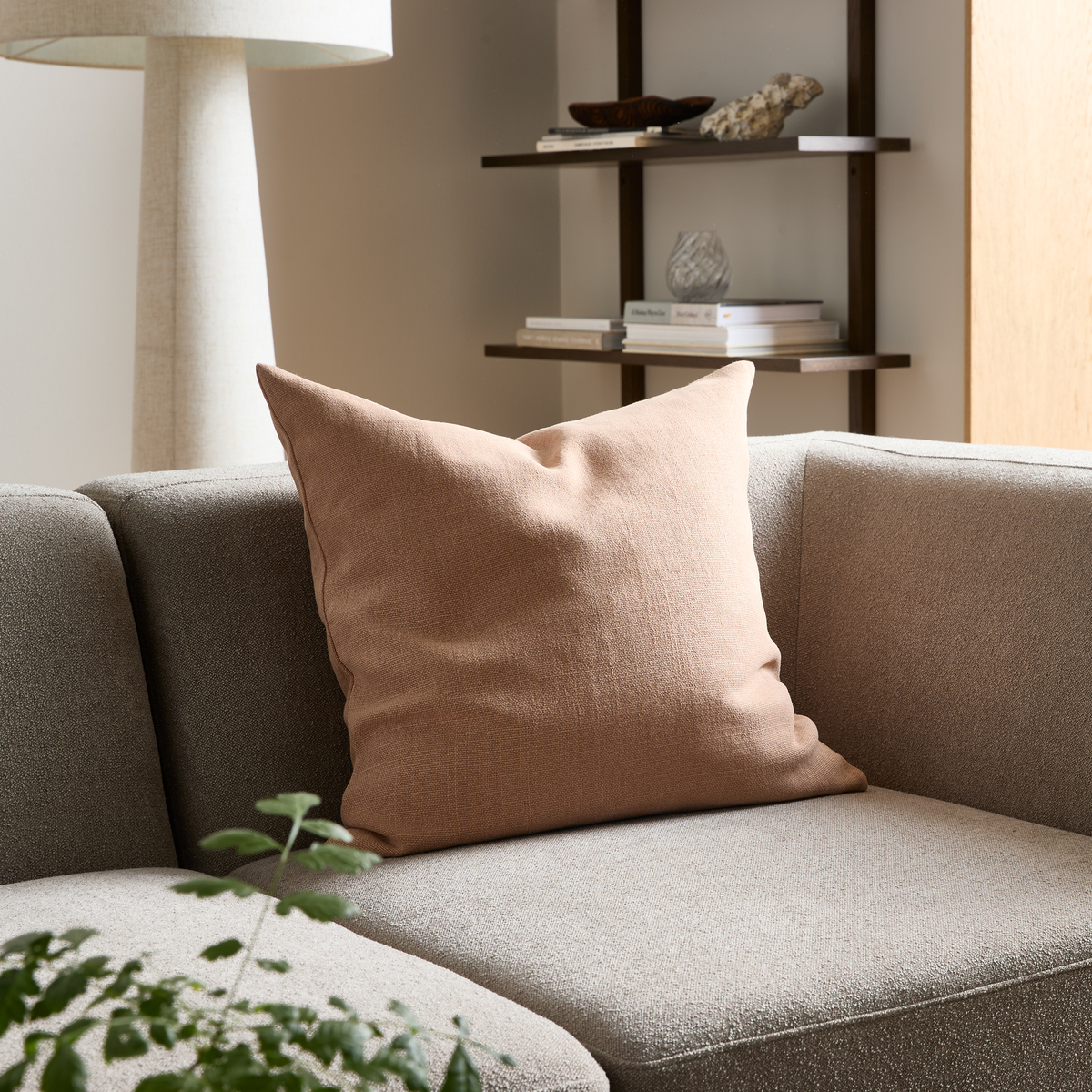 Beige pude på grå sofa i lys stue med gulvlampe, hylder med bøger og dekoration samt grøn plante i forgrunden.