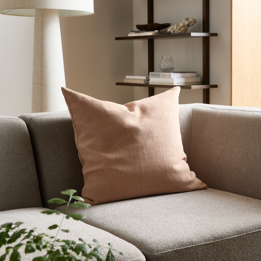 Beige pude på grå sofa i lys stue med gulvlampe, hylder med bøger og dekoration samt grøn plante i forgrunden.