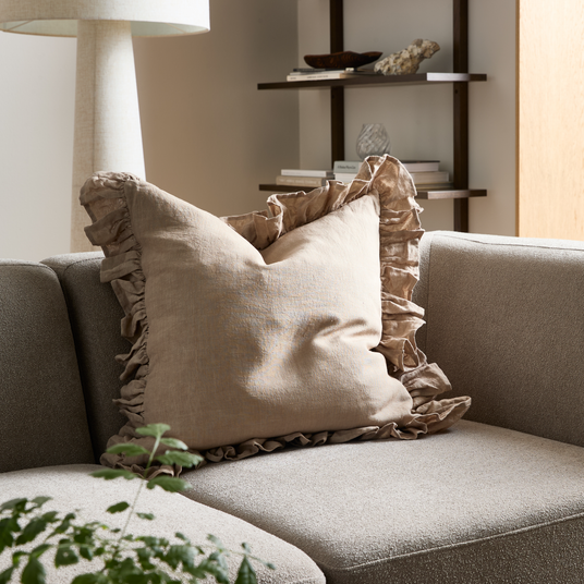 Beige pude med flæsekant på en grå sofa i en lys stue med gulvlampe, hylder og grøn plante.