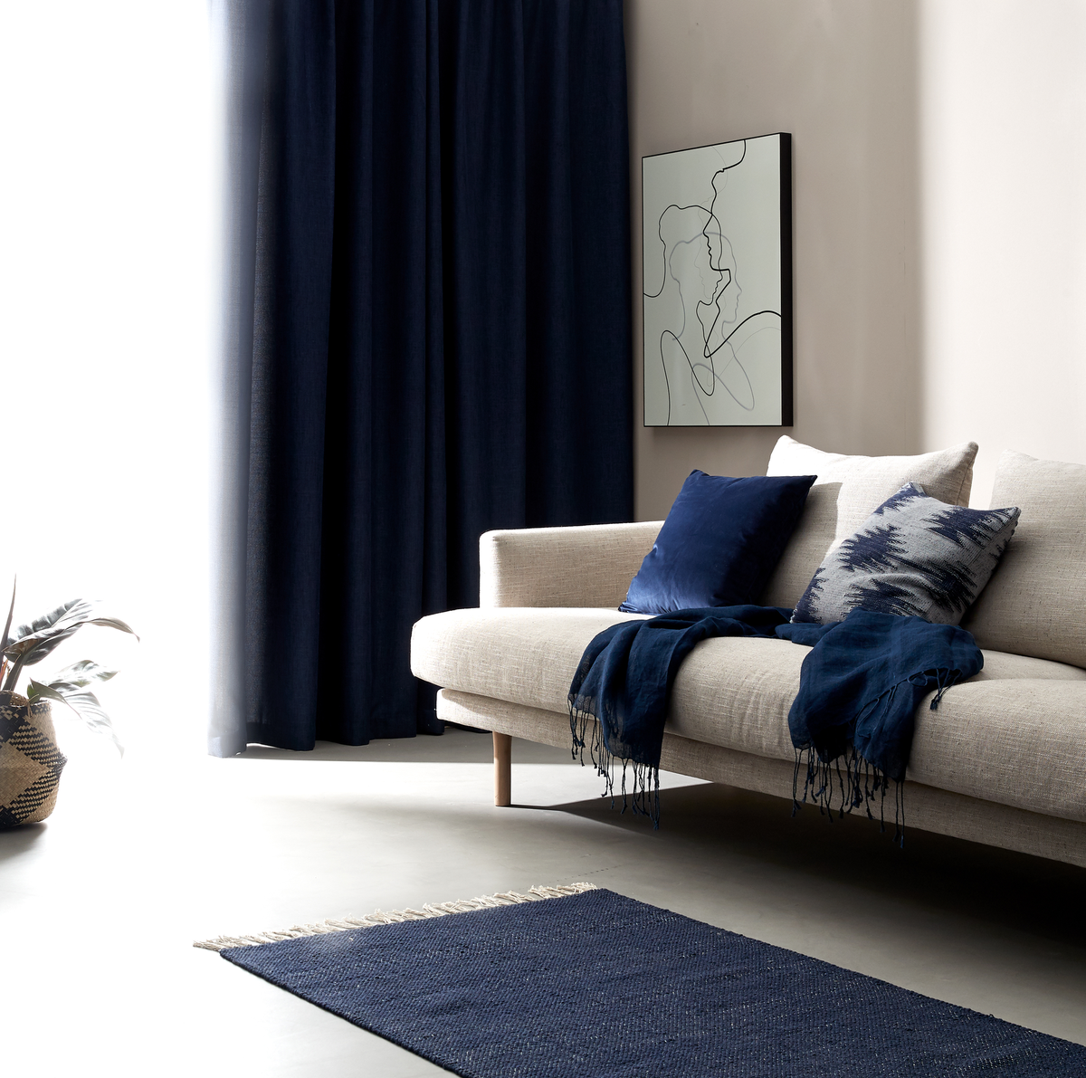 Beige sofa med mørkeblå puder og plaid, mørkeblå gardiner, blå løber og en plante i kurv i en lys, minimalistisk stue med lineart på væggen.