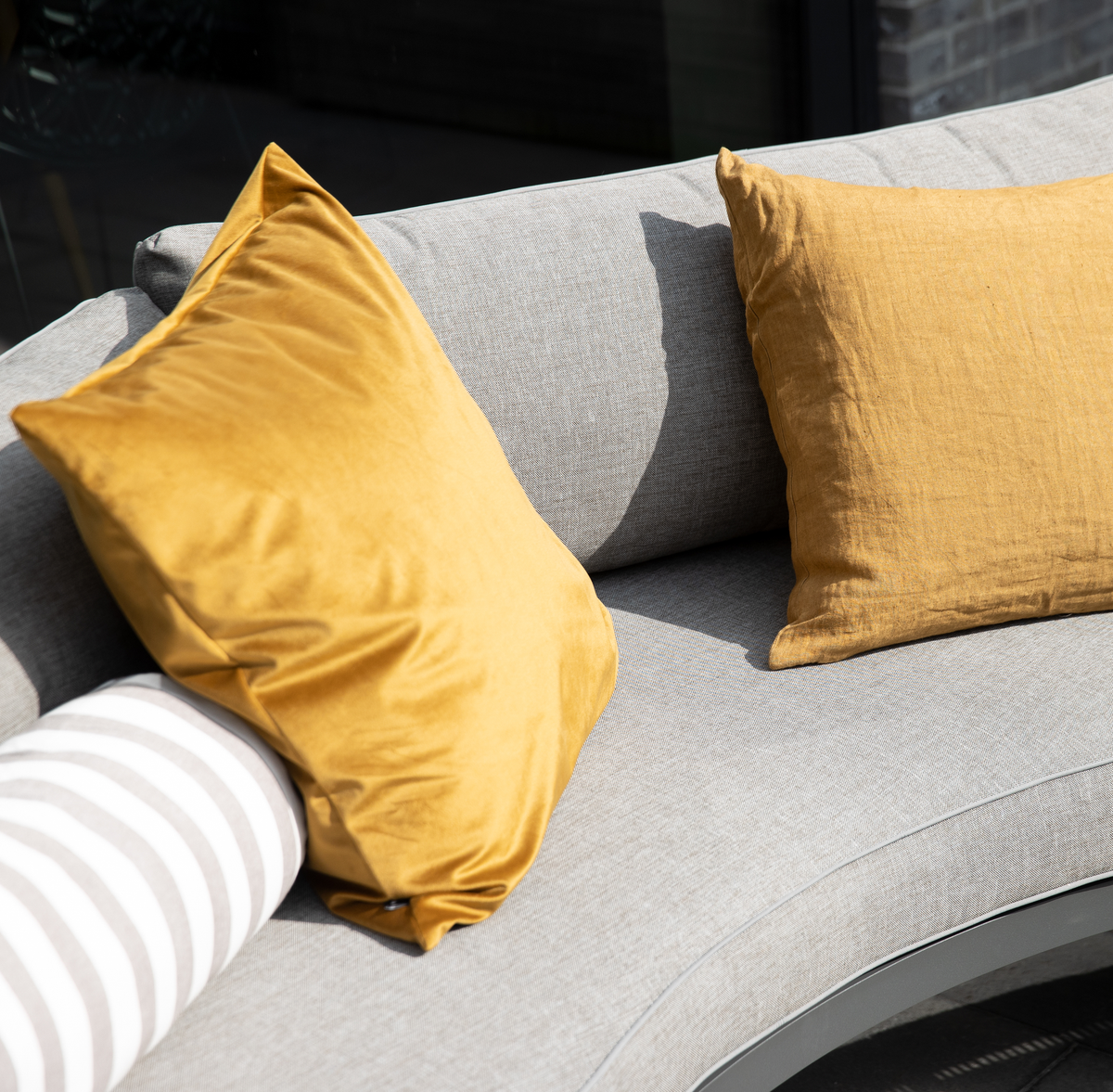 Grå sofa med to sennepsgule puder og en stribet bolster i sollys udenfor.