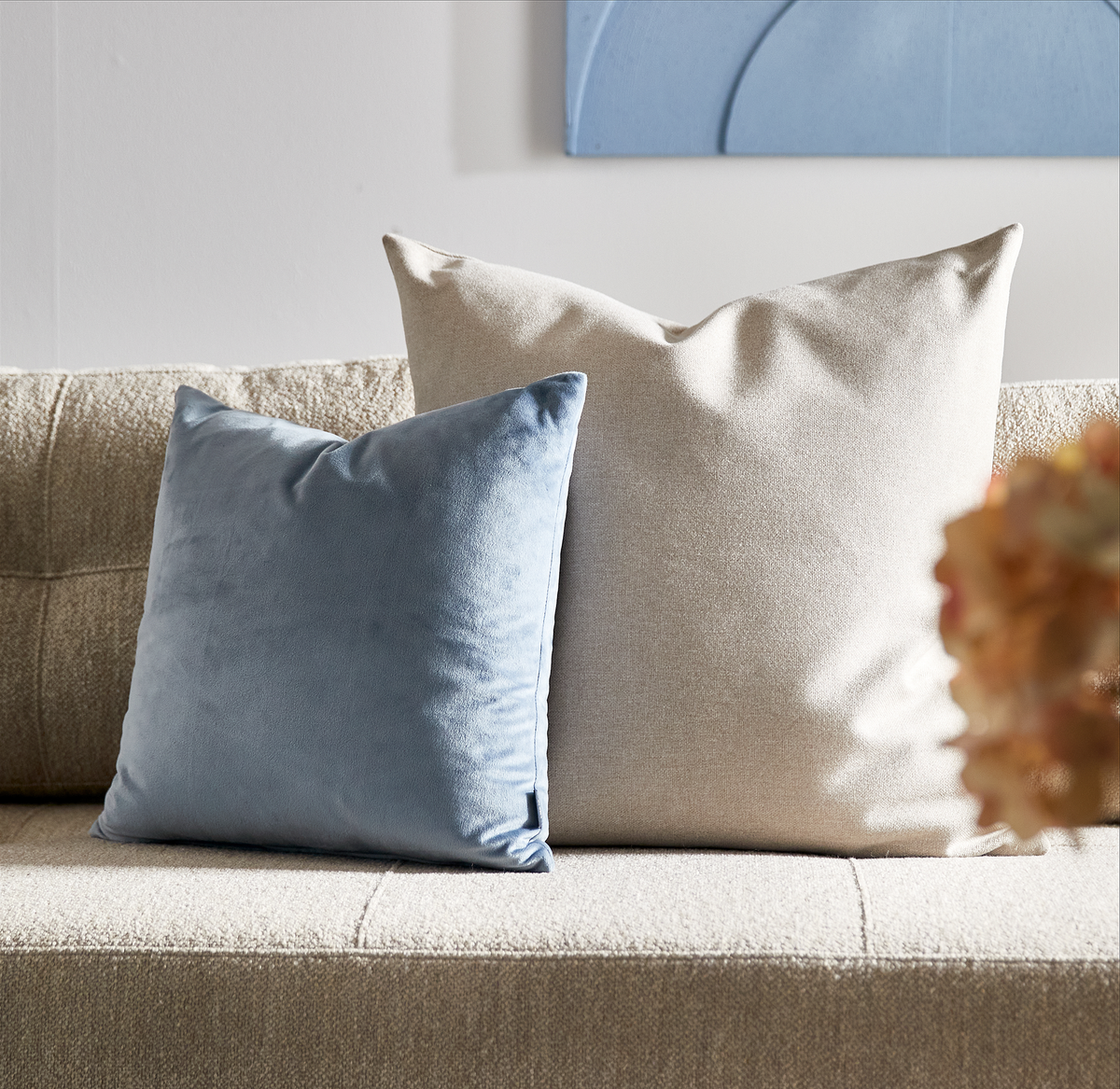 To pyntepuder på beige sofa: en større lysebeige og en mindre lyseblå velourpude.
