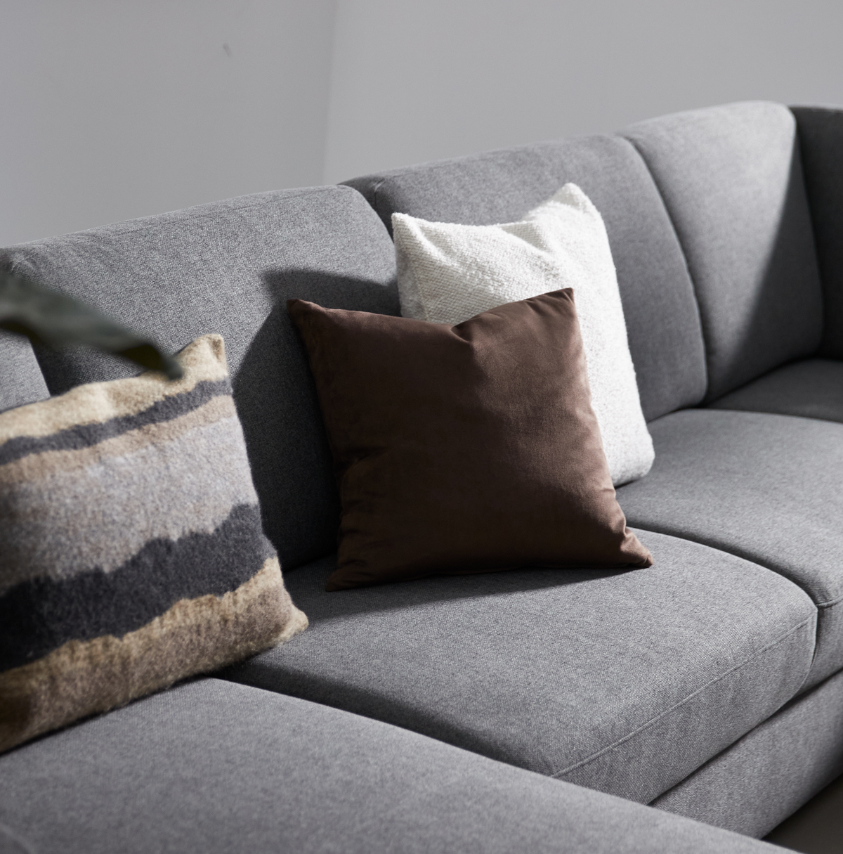 Grå sofa med tre pyntepuder – brun, hvid og mønstret i beige/grå nuancer.
