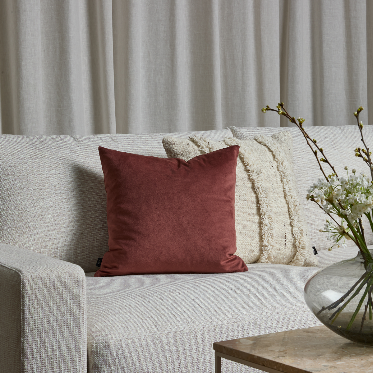 Beige sofa med rustfarvet pyntepude, lyse strukturpuder bagved og glasvase med hvide blomster på sofabord foran lyse gardiner.