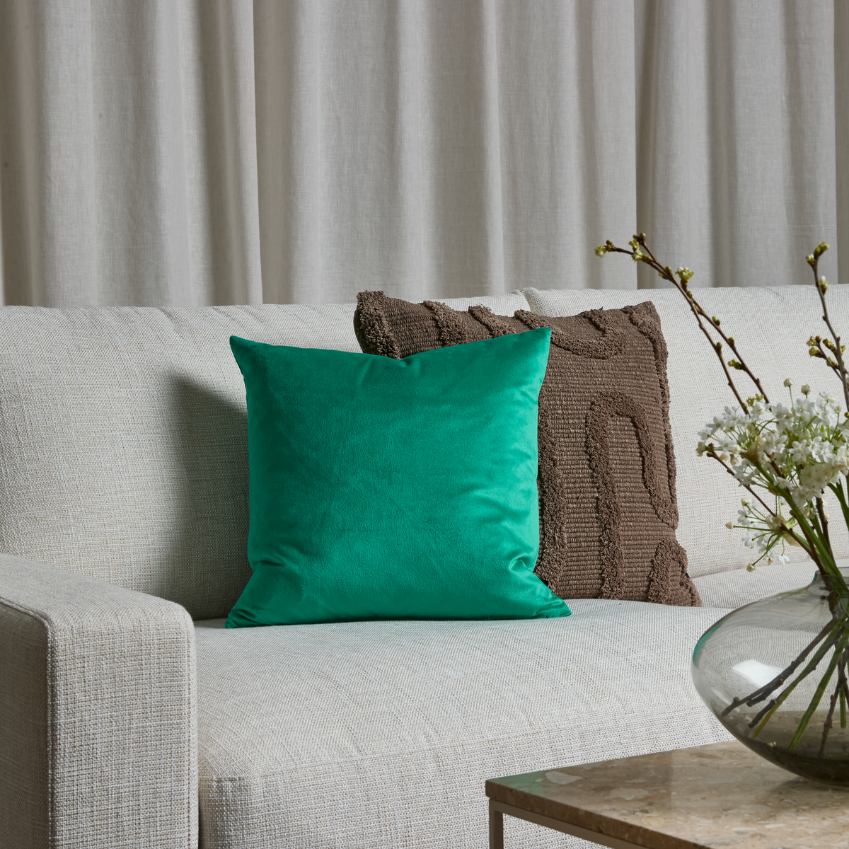 Lys sofa med grøn velourpude og brun struktureret pude; glasvase med hvide blomster på bord, lyse gardiner i baggrunden.